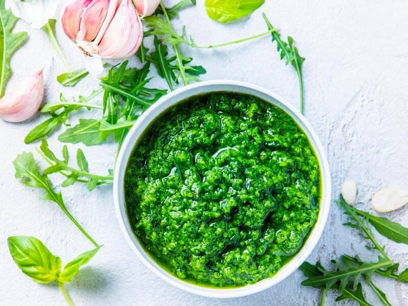 Bol blanc rempli de pesto vert frais avec ail, basilic et roquette autour sur fond clair.
