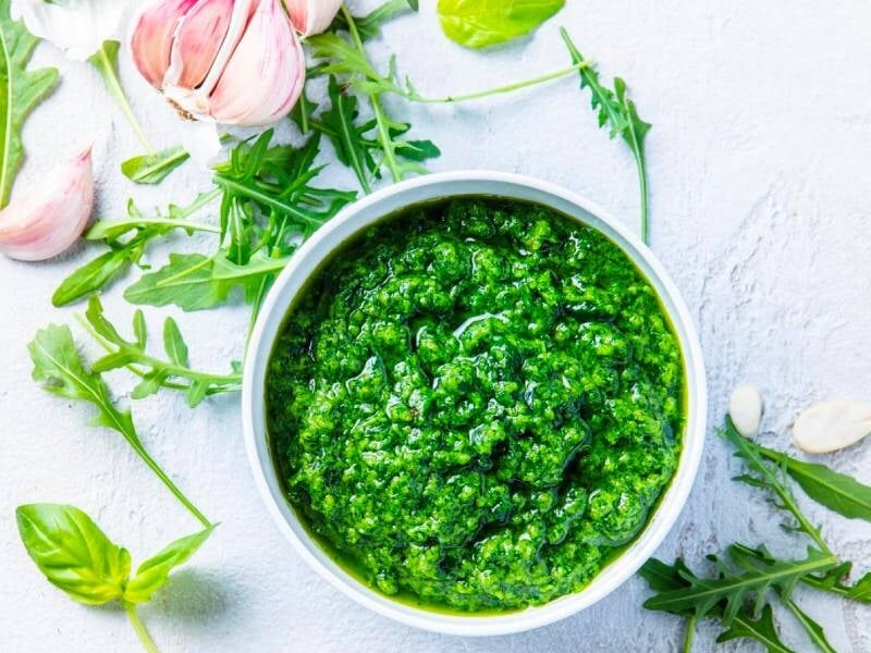 Bol blanc rempli de pesto vert frais avec ail, basilic et roquette autour sur fond clair.