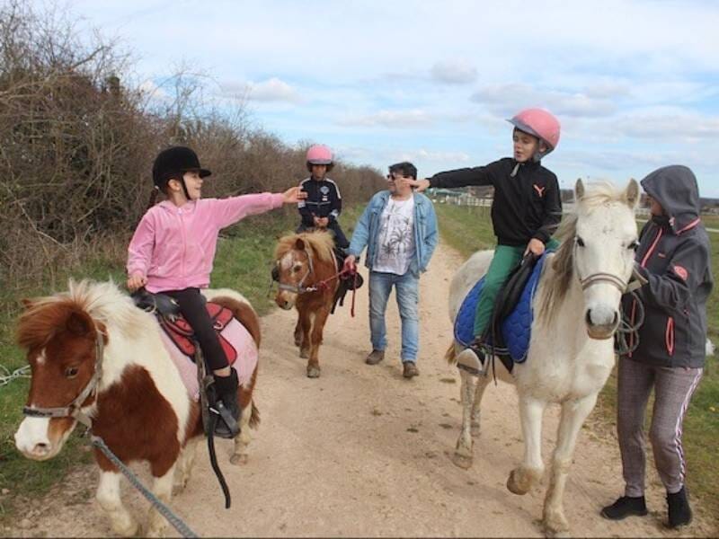 Enfants à cheval accompagnés d'adultes marchant sur un chemin de terre entouré de buissons et d'arbres.