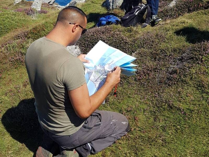 Homme agenouillé sur un sol herbeux, étudiant une carte lors d'une activité en plein air sous un ciel clair.