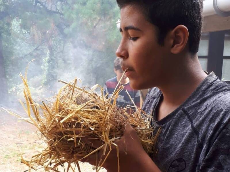 Jeune homme soufflant sur un nid de paille pour allumer un feu dans un environnement forestier brumeux, lors d'un stage de survie au Cap Sizun.