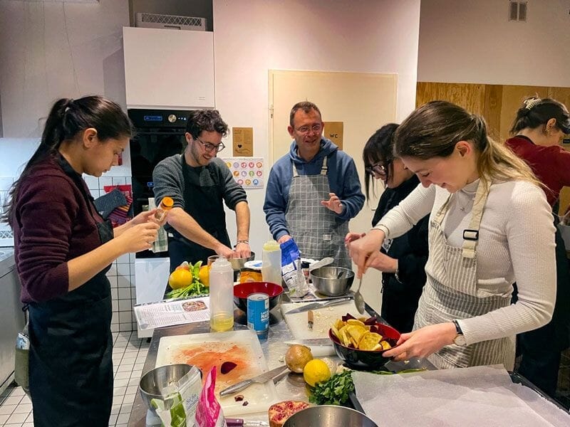 Groupe de personnes en tabliers cuisinant ensemble dans une cuisine moderne, échangeant sourires et techniques culinaires.