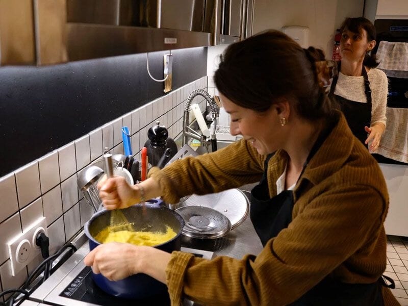 Jeune femme souriante mélangeant un plat dans une casserole sur une plaque de cuisson dans une cuisine équipée.