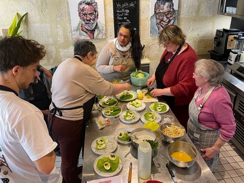 Groupe de personnes âgées et jeunes préparant des plats variés dans une cuisine avec des murs en pierre.