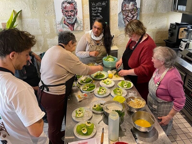Groupe de personnes âgées et jeunes préparant des plats variés dans une cuisine avec des murs en pierre.
