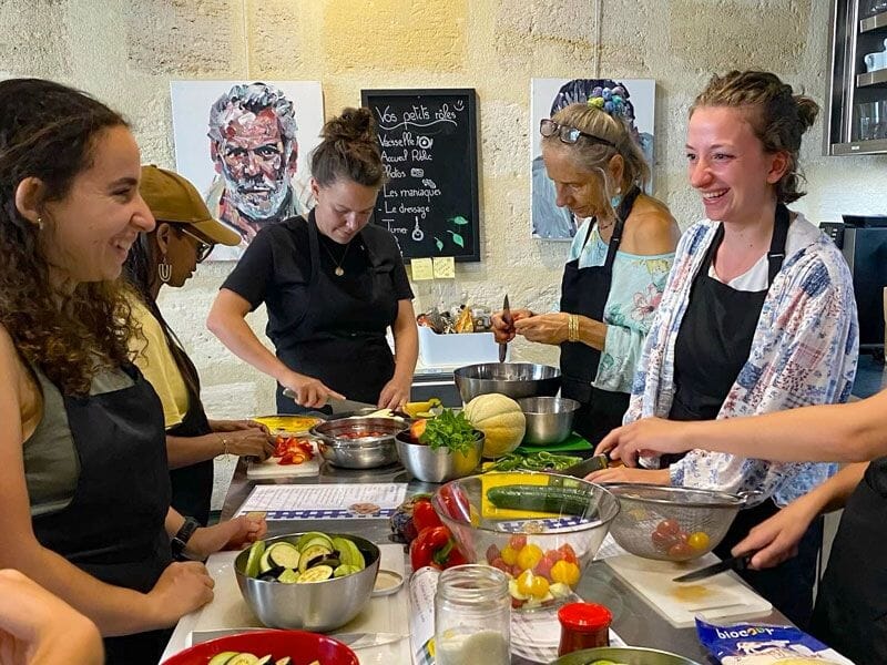 Groupe de femmes souriantes coupant des légumes frais et préparant des recettes dans une cuisine conviviale.
