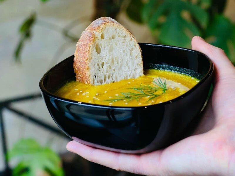 Bol noir contenant une soupe orange avec une tranche de pain posée sur le bord, présenté à la main.