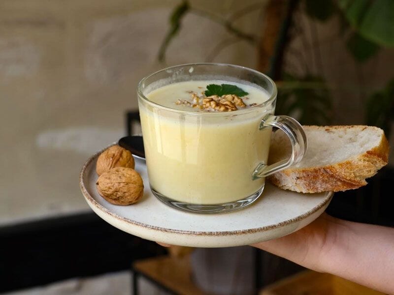 Verre transparent rempli d'une boisson crémeuse, accompagné de noix et d'une tranche de pain sur une assiette.