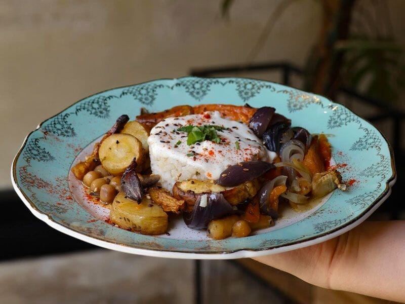 Assiette colorée avec un plat composé de légumes rôtis, pois chiches et un œuf poché, tenue à la main.