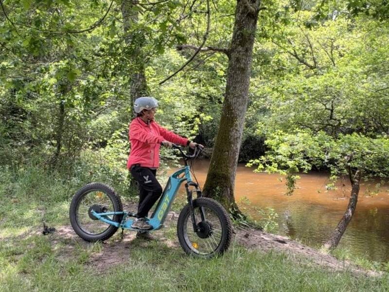 Personne en tenue rouge et casque gris sur une trottinette électrique bleue près d'un cours d'eau en forêt, lors d'une randonnée près de Langon.