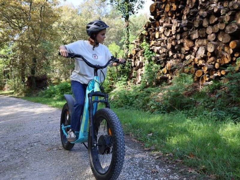 Jeune femme en casque noir sur une trottinette électrique bleue sur un chemin forestier à côté d'un tas de bois.