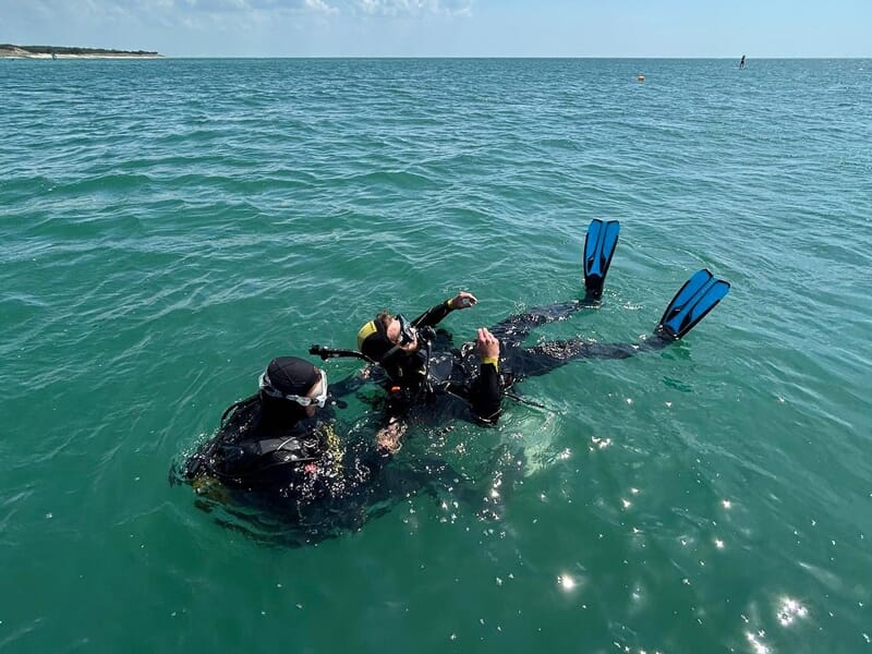 Deux plongeurs en combinaison noire flottent à la surface de l'eau, l'un aidant l'autre lors d'un baptême de plongée.