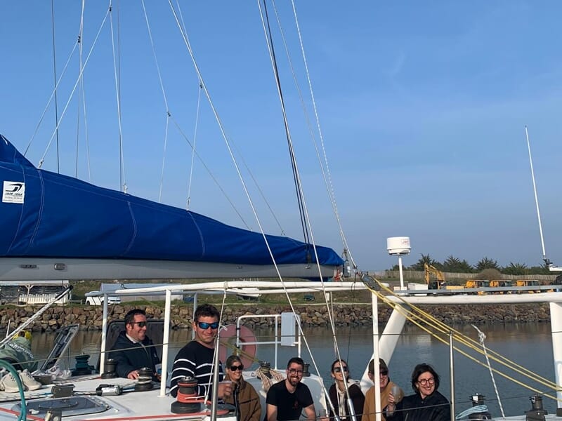 Un groupe de personnes souriantes à bord d'un voilier amarré dans un port avec un ciel bleu en arrière-plan.