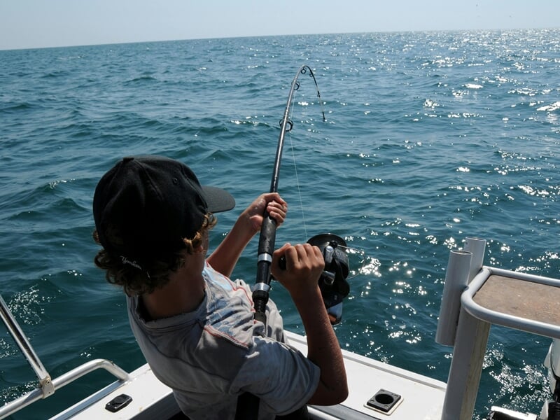 Personne en train de pêcher au gros depuis un bateau sur l'océan, avec une canne à pêche courbée par la tension.