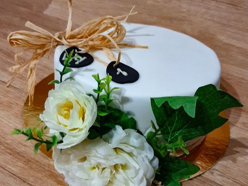 Gâteau blanc décoré avec fleurs blanches et feuilles vertes sur table en bois clair, ambiance naturelle.
