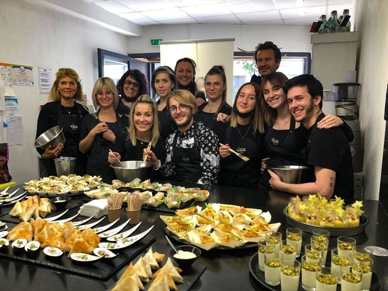 Groupe de personnes souriantes en tabliers noirs posant derrière une table garnie de plats variés et appétissants.
