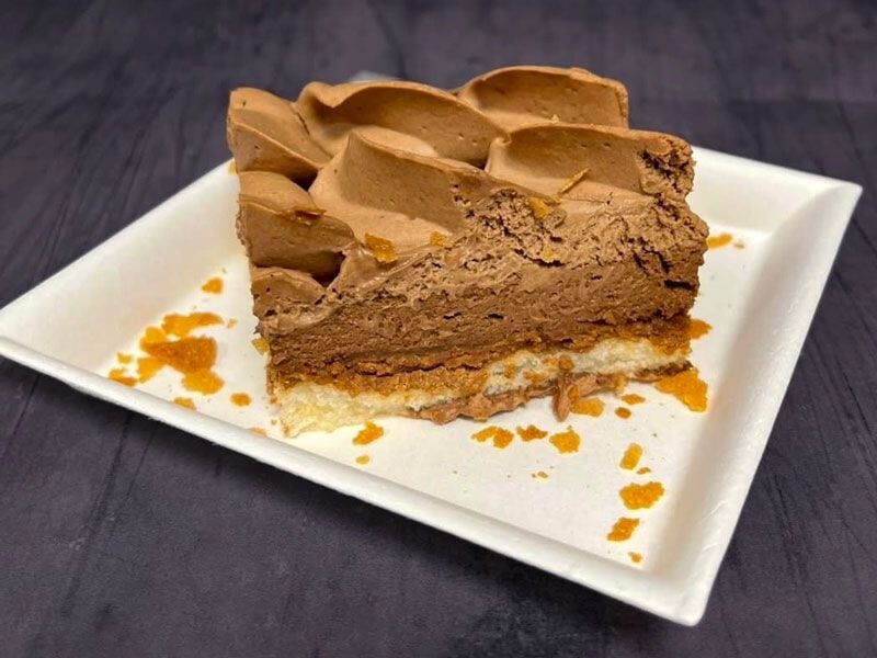 Part de gâteau au chocolat mousseux sur assiette blanche avec miettes dorées autour, fond sombre.