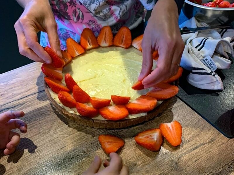 Mains disposant des fraises coupées en quartiers sur une tarte à la crème sur planche en bois.