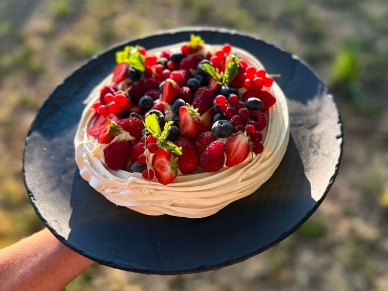 Pavlova décorée de fruits rouges frais et feuilles de menthe, tenue à la main sur assiette noire.