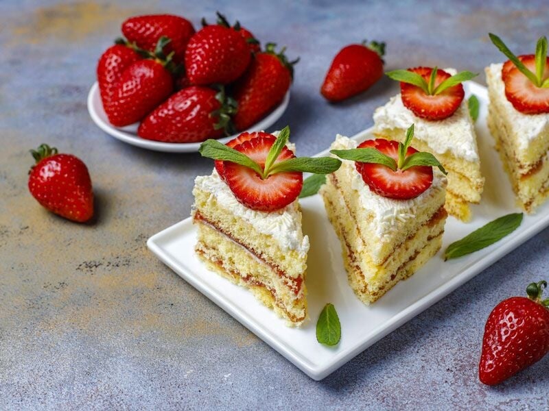 Petits gâteaux à la crème garnis de fraises et feuilles de menthe, avec fraises entières en arrière-plan.