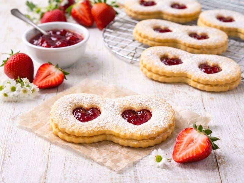 Biscuits sablés en forme de huit avec confiture rouge au centre, décorés de sucre glace et fraises fraîches à côté.