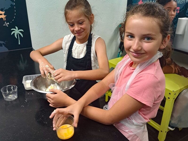 Deux jeunes filles souriantes en tablier préparent une recette de cuisine ensemble dans une ambiance conviviale.