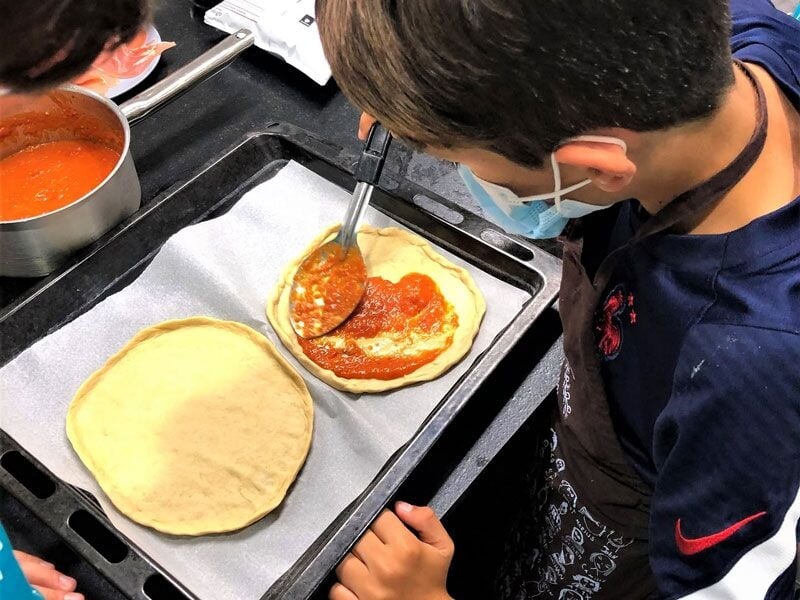 Un enfant étale de la sauce tomate sur une pâte à pizza dans un cours de cuisine pour enfants.