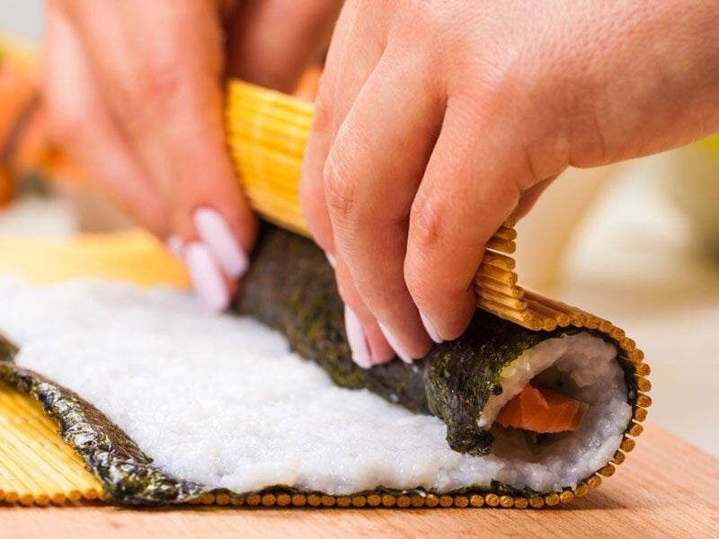 Mains en train de rouler une feuille d'algue avec du riz et du saumon pour préparer un sushi maison.