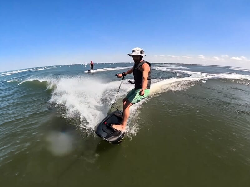 Homme en short vert et chapeau blanc surfant sur une planche électrique dans une mer agitée.