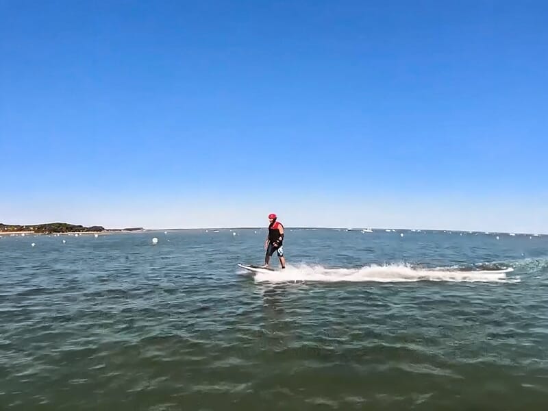 Surfeur en combinaison noire et casque rouge sur une planche électrique sur une mer calme avec des bouées.