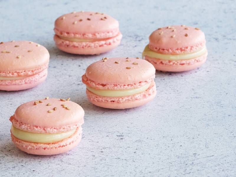 Cinq macarons roses garnis d'une crème blanche, posés sur une surface claire avec des éclats dorés.