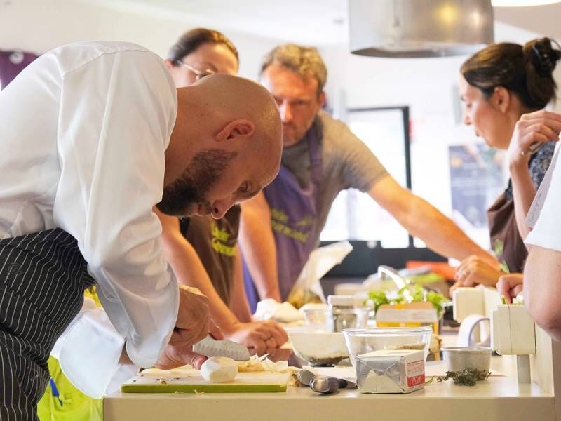 Chef concentré préparant un plat avec des ingrédients frais, entouré de participants attentifs dans un atelier culinaire.