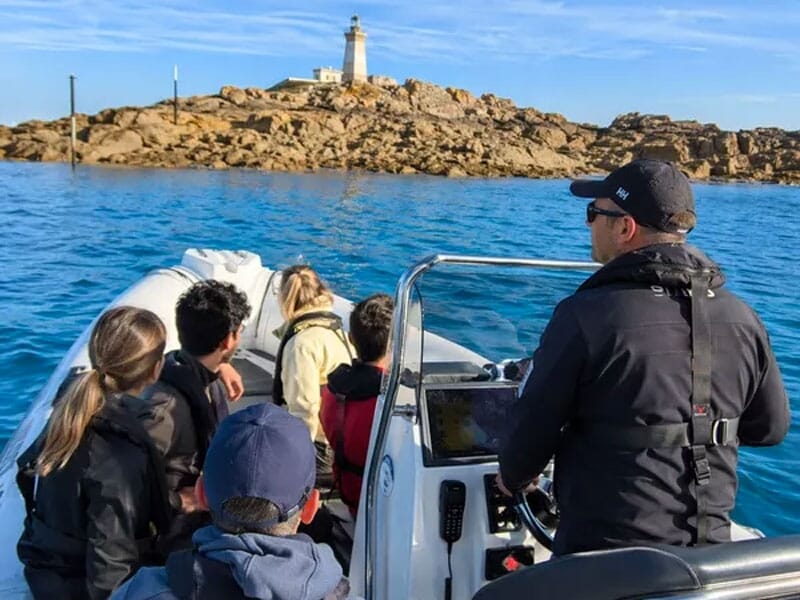 Groupe de personnes en bateau se dirigeant vers un phare sur une île rocheuse sous un ciel bleu clair.