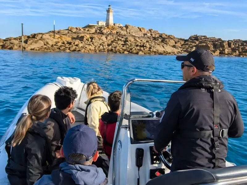 Groupe de personnes en bateau se dirigeant vers un phare sur une île rocheuse sous un ciel bleu clair.