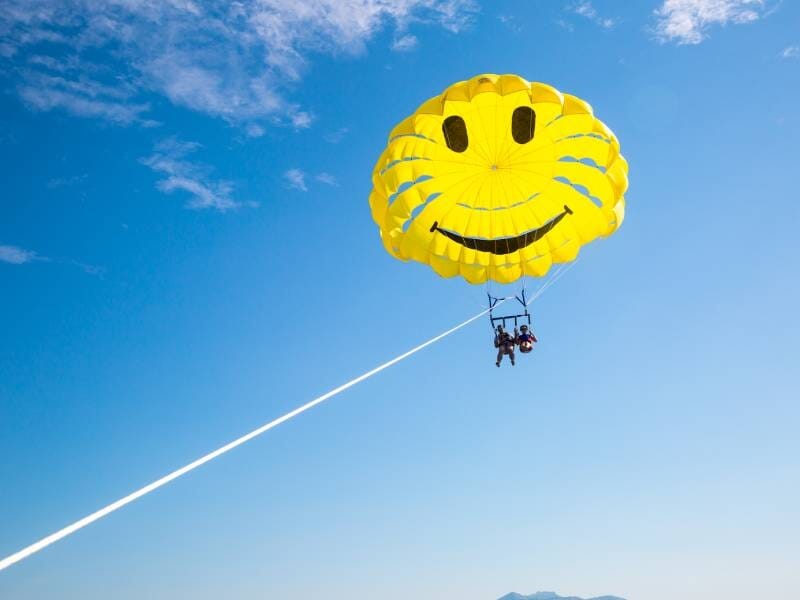 Deux personnes en parachute ascensionnel sous une voile jaune souriante, tirées par un bateau sur une mer calme.