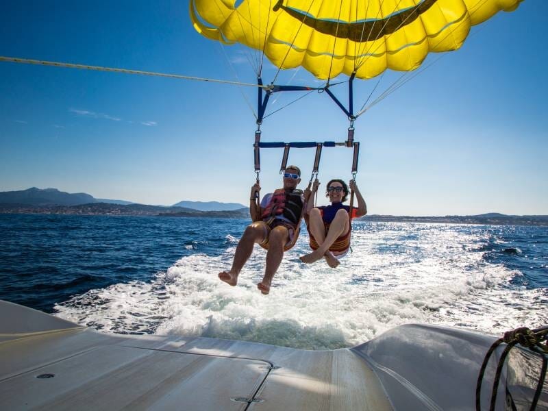 Couple en parachute ascensionnel souriant, suspendu au-dessus de la mer, avec un bateau blanc en arrière-plan, lors d'un vol à Saint-Cyr-sur-Mer.