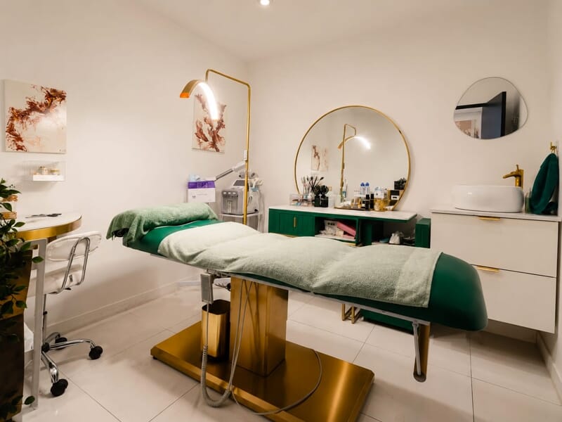 Salle de soin avec table de massage, miroir rond et mobilier vert dans un cadre élégant et propre.