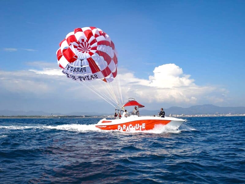 Bateau orange et blanc tirant un parachute ascensionnel rouge et blanc avec des passagers à bord.