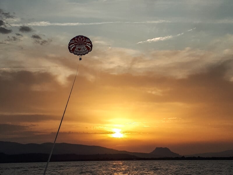 Parachute ascensionnel au coucher du soleil avec un ciel orangé et des montagnes en arrière-plan sur l'eau.