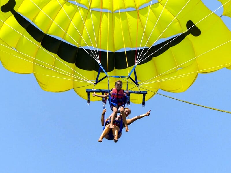 Deux personnes en parachute ascensionnel sous un parachute jaune, suspendues au-dessus de la mer avec un ciel bleu.