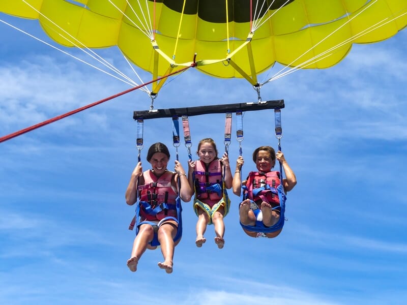 Trois personnes en parachute ascensionnel avec un parachute jaune souriant au-dessus de la mer et ciel bleu clair.
