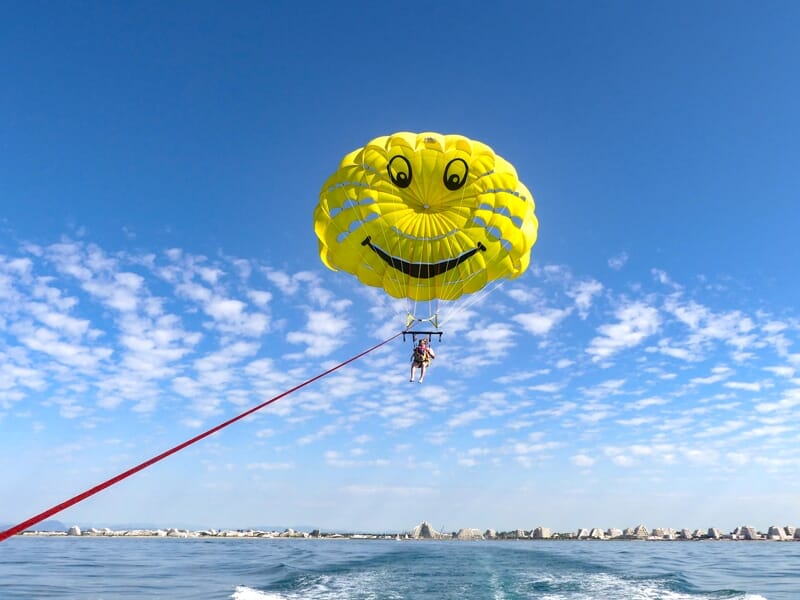 Parachute ascensionnel jaune avec un visage souriant, deux personnes suspendues au-dessus de la mer calme et ciel bleu.