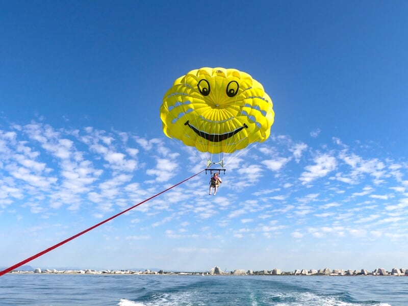 Parachute ascensionnel jaune avec un visage souriant, deux personnes suspendues au-dessus de la mer calme et ciel bleu.