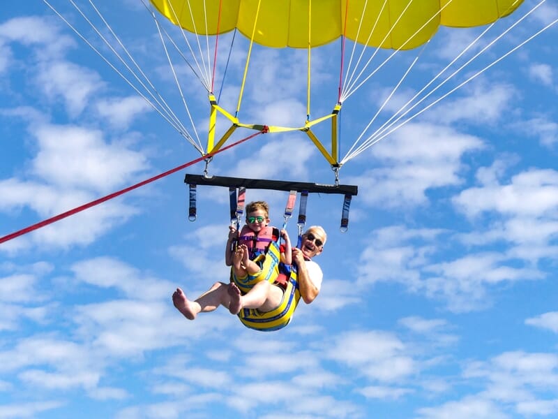 Un adulte et un enfant en parachute ascensionnel avec un parachute jaune, ciel bleu et nuages blancs en arrière-plan.