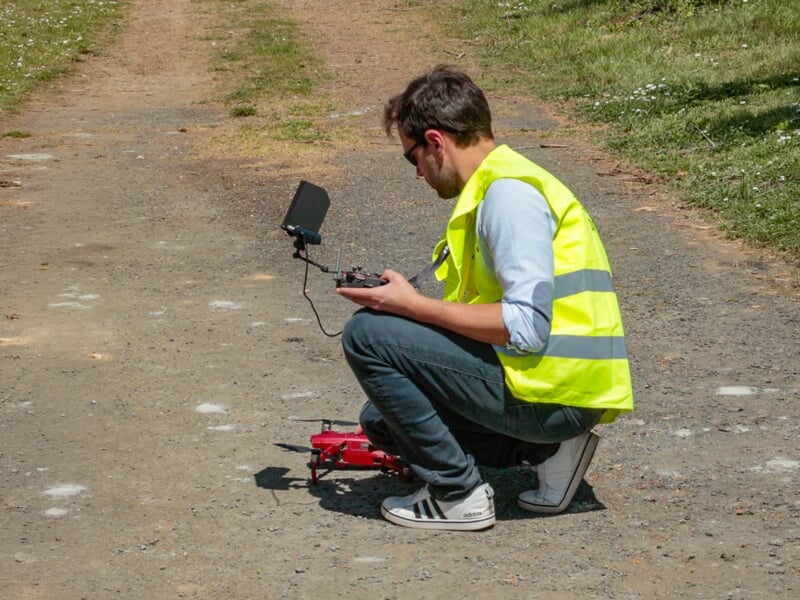 Homme accroupi en gilet jaune pilotant un drone rouge avec une télécommande et un écran sur un chemin en terre.