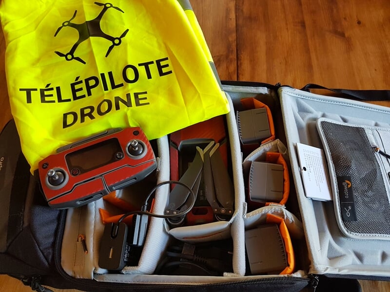 Sac ouvert contenant un drone, une télécommande rouge et un gilet jaune avec l'inscription Télépilote Drone posé sur une table en bois.