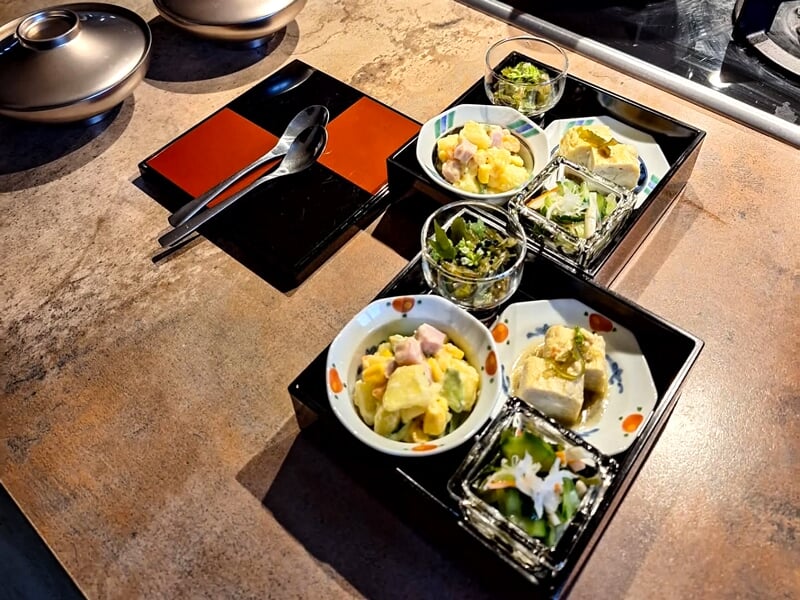 Assortiment de petites entrées japonaises servies dans des bols et plateaux noirs sur une table en bois.