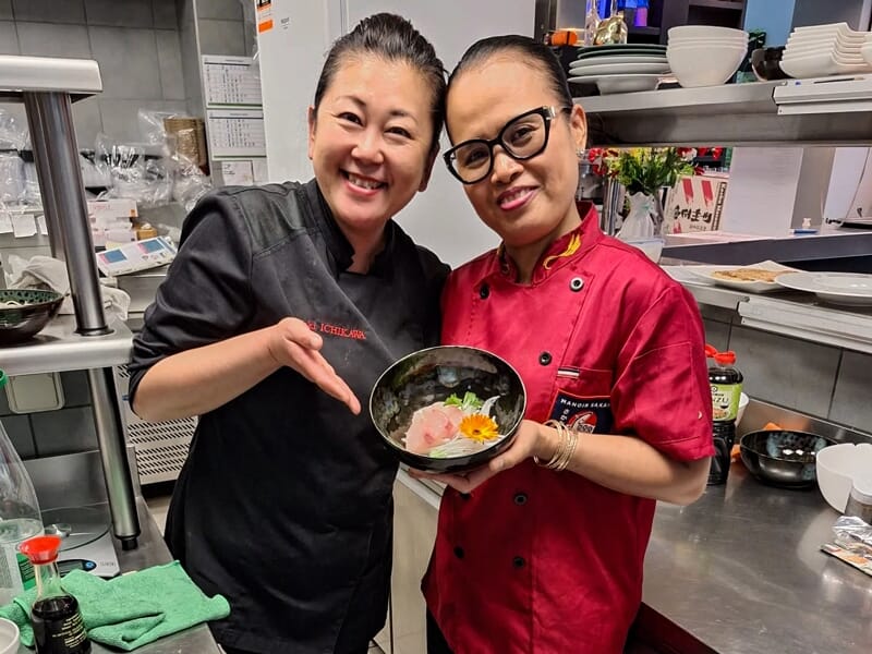 Deux cuisinières souriantes présentent un plat japonais préparé dans une cuisine professionnelle.