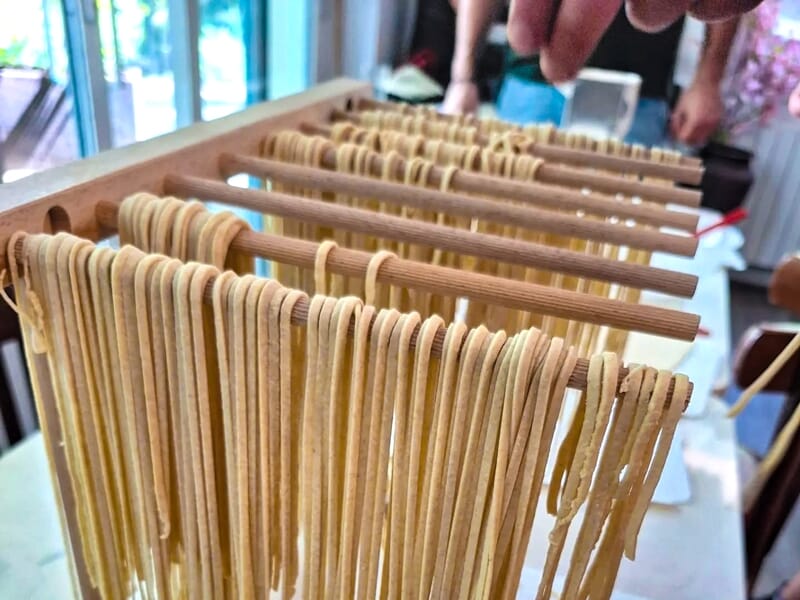 Pâtes fraîches étirées et suspendues sur des bâtons en bois pour sécher dans une cuisine lumineuse.
