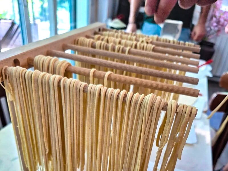 Pâtes fraîches étirées et suspendues sur des bâtons en bois pour sécher dans une cuisine lumineuse.